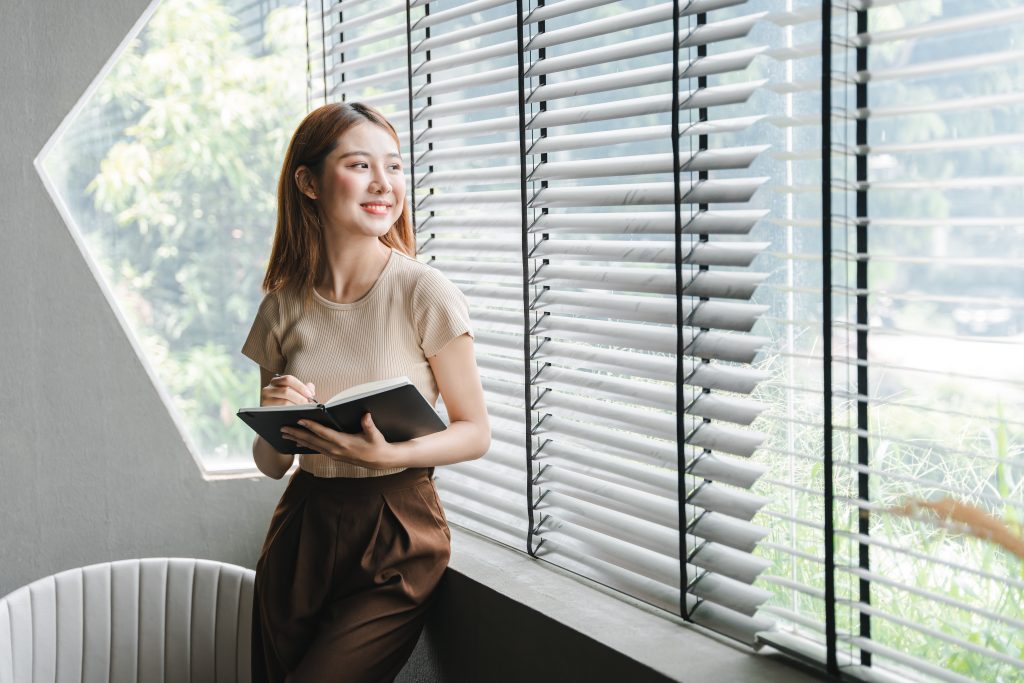 Smiling Asian woman holding a note in hand and standing near window with blinds