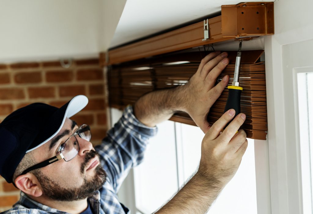 Man with mustache wearing cap and eyeglass installing window blinds